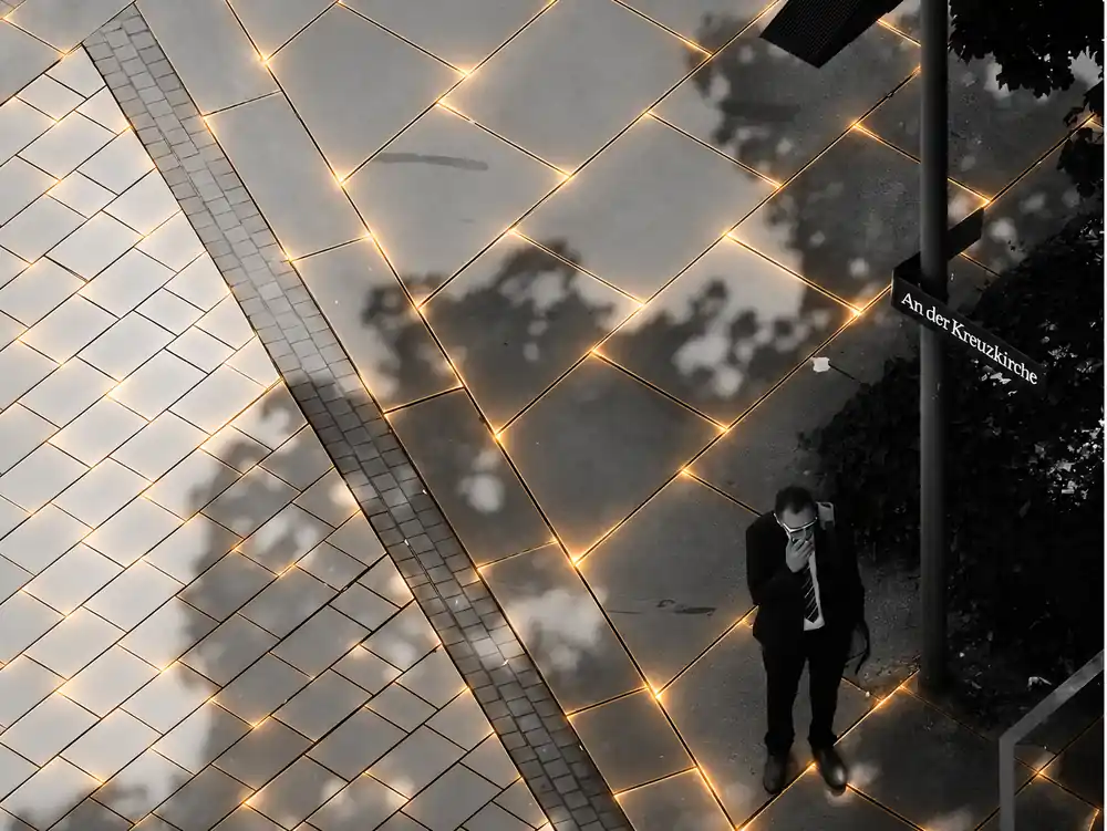 Acera con baldosas solares inteligentes Suntile de Deca: juntas iluminadas en tono dorado y peatón caminando junto a una farola